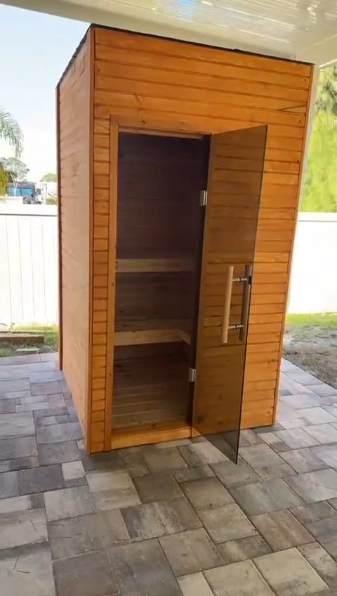 Compact amber-stained sauna cabin on a paver patio, its full-height tinted glass door open to two stepped benches