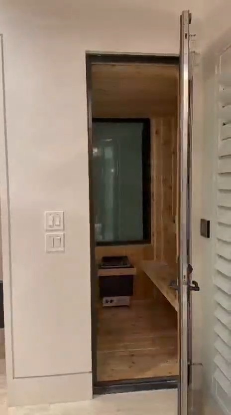 Black-framed glass sauna door open in a white bathroom wall, showing wood walls, a window, heater and bench inside