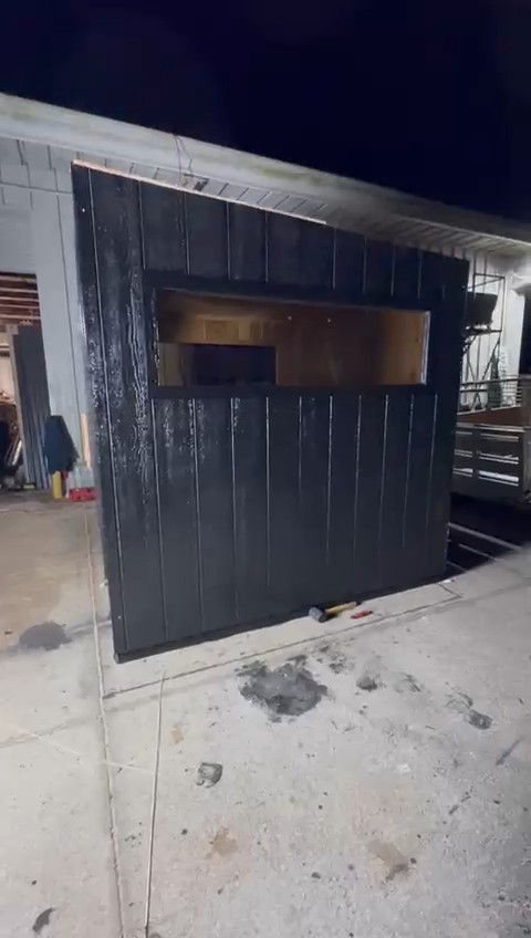 Black vertical-board cabin sauna at night beside a metal workshop, with a long window high on its side wall