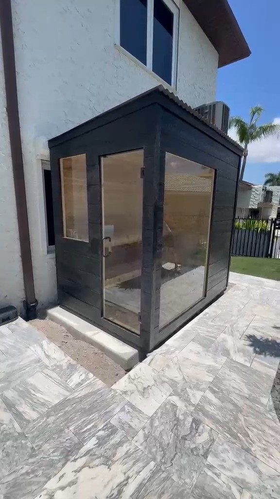 Black-clad cube sauna with a small window, glass door and tall side glass panel beside a white stucco house on a paved patio