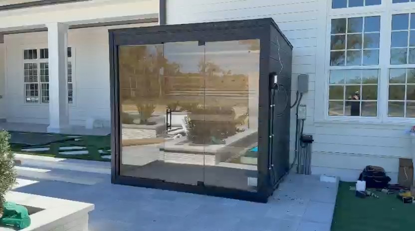 Black-framed cube sauna with a full-height glass front reflecting the yard, on a grey patio beside a white house