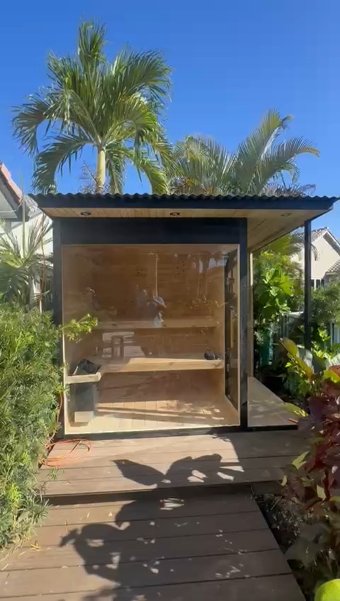 Black-framed outdoor sauna with a full glass front and corrugated metal roof on a dark deck, palm trees behind