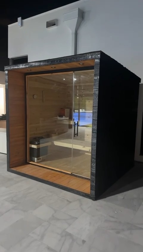 Black-clad outdoor sauna with a full glass front and a wood-lined, downlit portal on a tiled poolside patio at night