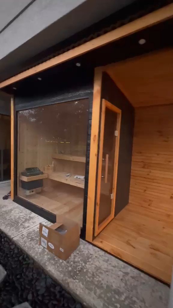 Dark-clad sauna with a black-framed glass front, a wood-framed glass door and a covered porch lined in knotty boards
