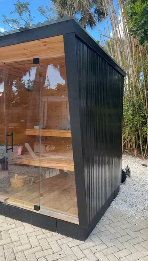 Black vertical-board outdoor sauna with a full-height glass front on a herringbone paver patio, bamboo behind