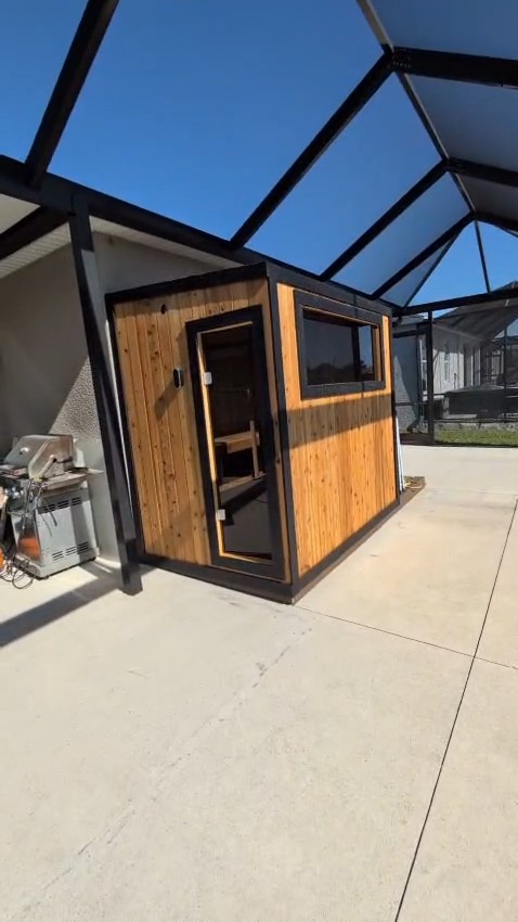 Rectangular sauna cabin in amber vertical boards with black trim, a glass door and side window on a screened concrete patio