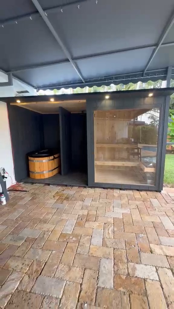 Charcoal-clad sauna module under a patio roof: open bay with wooden cold plunge tub, and a glass-front sauna on the right