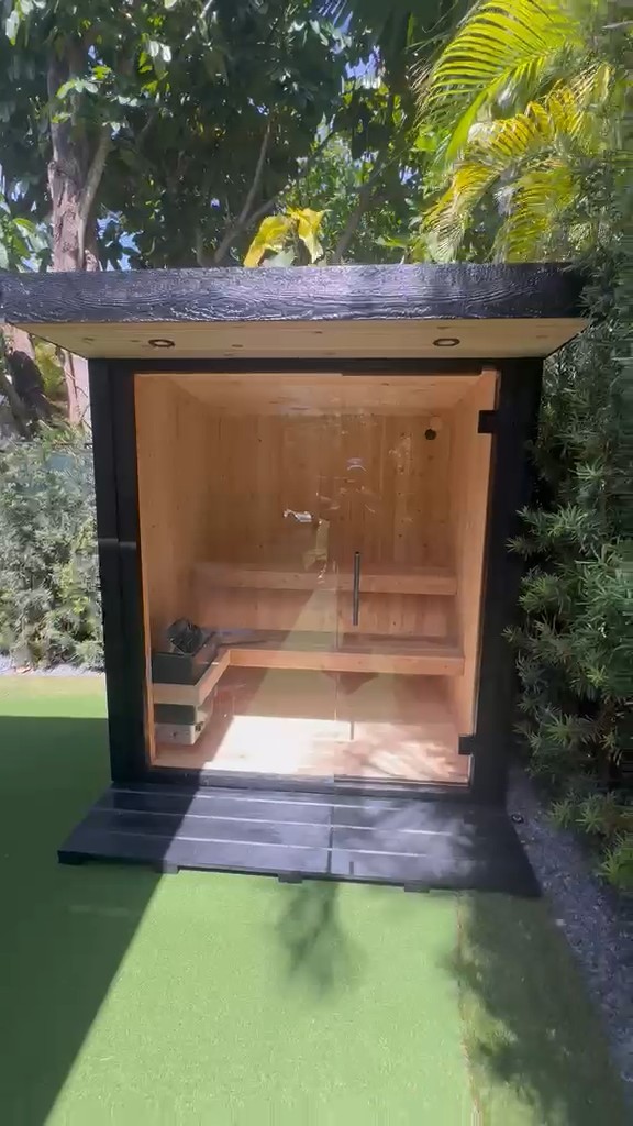 Front view of a compact black sauna with a full-glass front and pale wood soffit, on a black slatted pad on green turf
