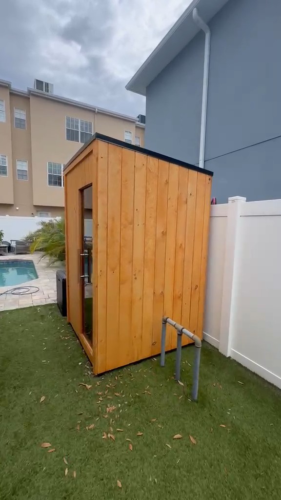Compact sauna clad in honey-finish vertical boards with a smoked glass door, on turf by a white fence and pool