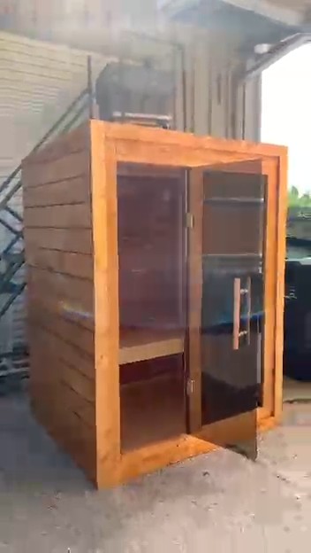 Compact cabinet sauna with its tinted glass door ajar, a wide amber-stained wood frame and horizontal board siding