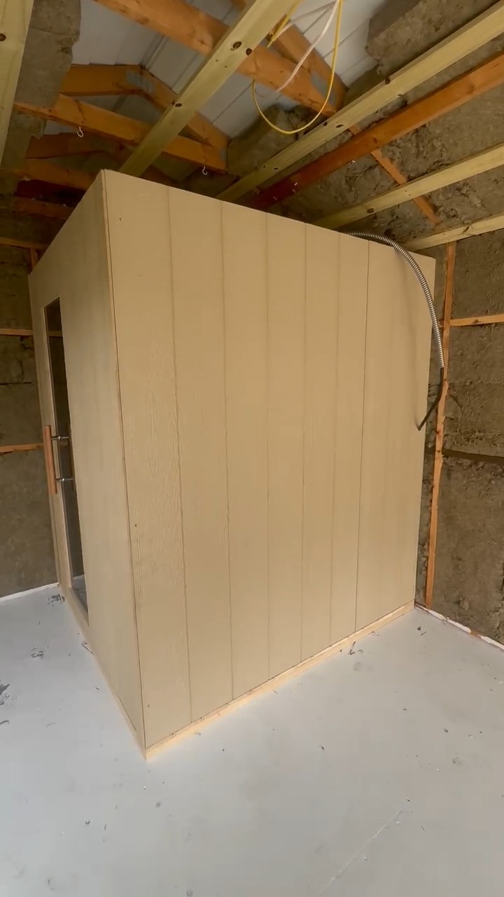 Freestanding indoor sauna in cream vertical panel siding with a smoked glass door, on a concrete floor under open rafters