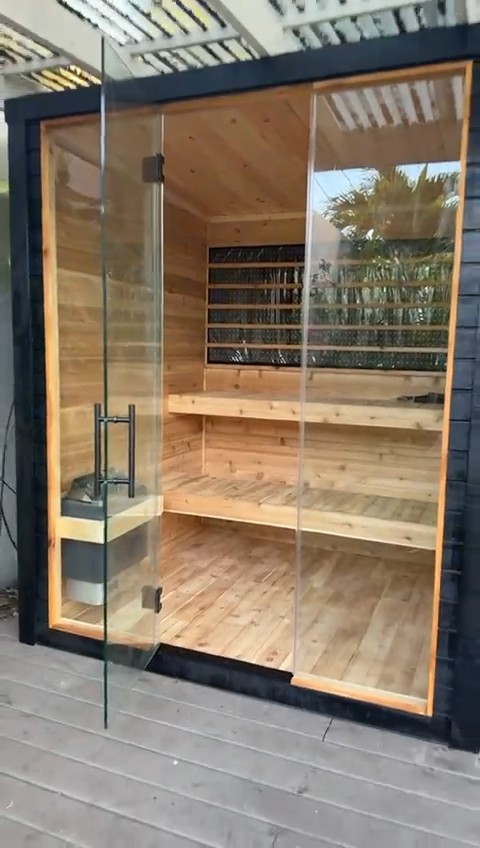 Open glass door of a black-clad sauna showing two slatted bench tiers, a rear window band and the stone heater