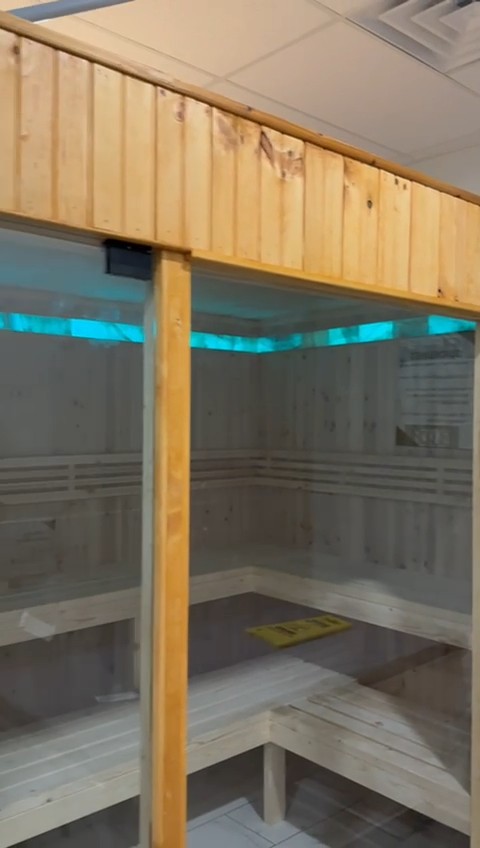 Glass front of an indoor sauna cabin under an amber-finished board header, with cyan LED and benches seen through the glass