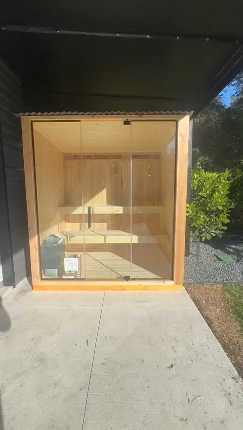 Compact sauna cabin with a full glass front and wood frame, on a concrete pad under a black patio overhang
