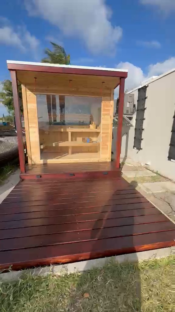 Glass-front outdoor sauna under a covered porch with dark red posts and a wide stained deck on a lawn