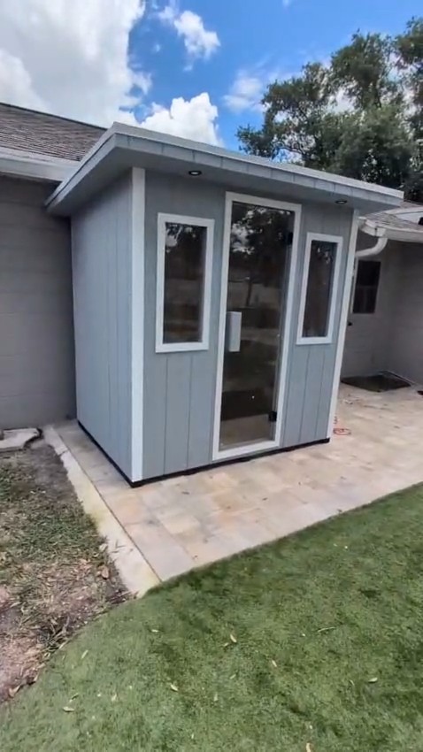 Grey-sided outdoor sauna cabin with a full-height glass door between two white-trimmed windows, on a paver patio by a house