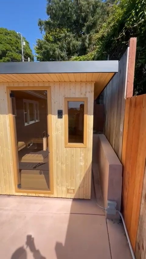 Pale vertical-board sauna with tinted full-height glass door, narrow window and black-edged roof overhang on a patio