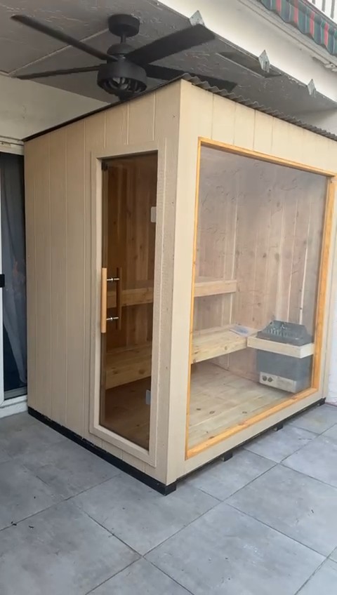 Compact sauna cabin in beige grooved siding with a glass end door and a wood-framed glass front on a covered tiled patio