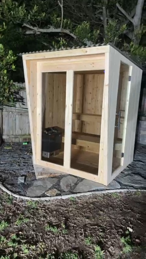 Outdoor sauna at night: two-panel glass front in a pale wood frame, corrugated shed roof, glass side door, flagstones