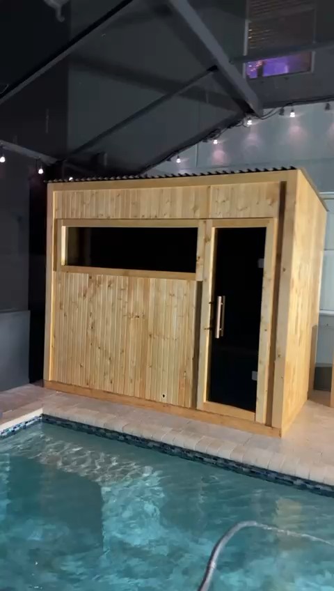 Pale vertical-board sauna cabin with a dark glass door and wide tinted window beside a pool in a screened enclosure at night