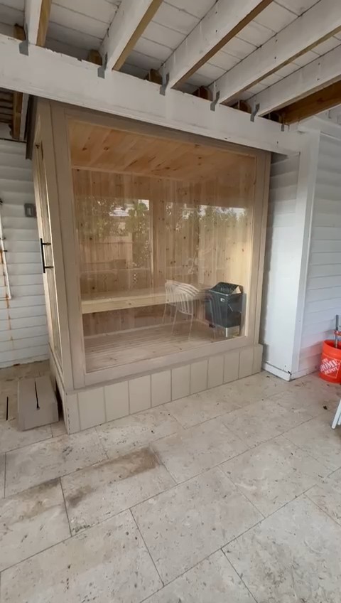 Sauna with a full-height glass front, cream frame and taupe board base, under exposed joists on a tile patio