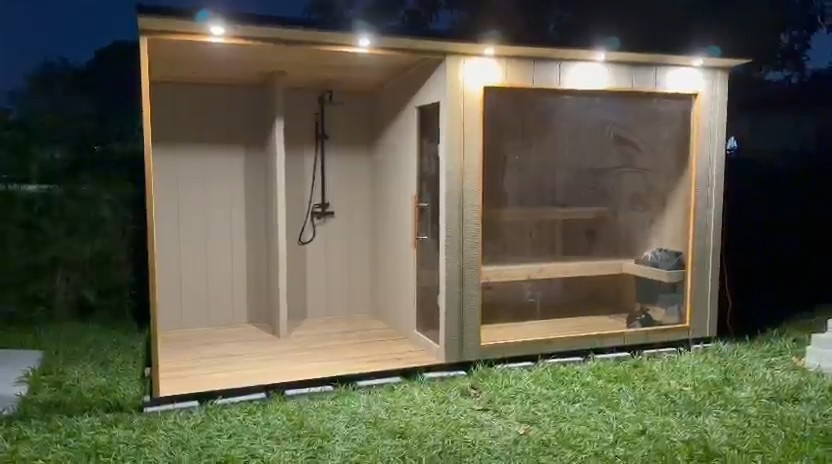 Two-bay outdoor sauna on a lawn at night: open shower bay on the left, glass-fronted sauna room on the right