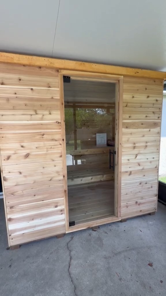 Rectangular sauna clad in horizontal knotty wood boards with a full-height glass door, standing on a covered patio slab