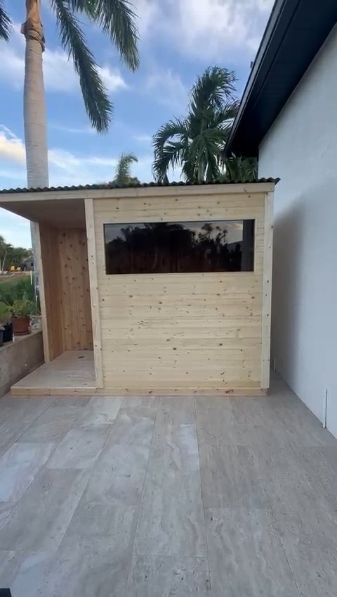 Pale knotty-board sauna with a wide tinted window, black corrugated shed roof and open side alcove on a tiled terrace