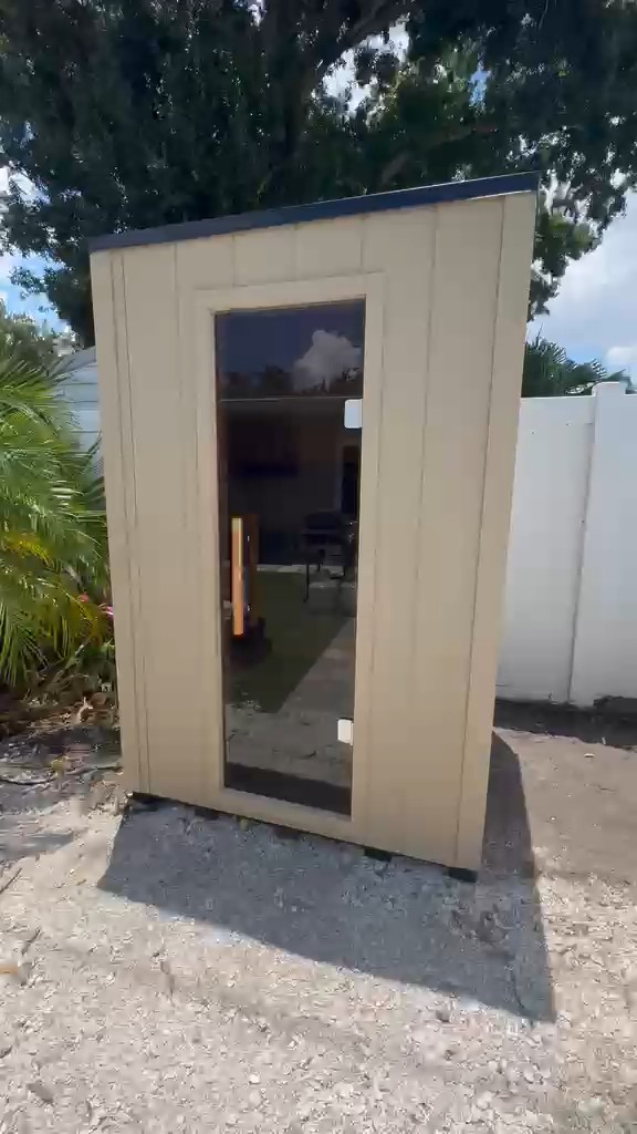 Compact outdoor sauna with tan vertical siding, dark roof edge and full-height glass door on a gravel pad by a white fence