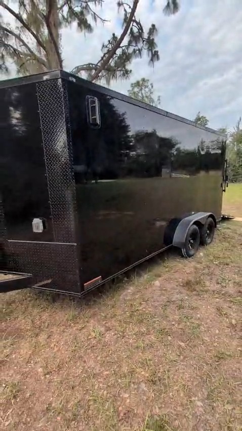 Gloss-black tandem-axle trailer sauna seen from the front corner, with diamond-plate trim, on a grassy lot under trees